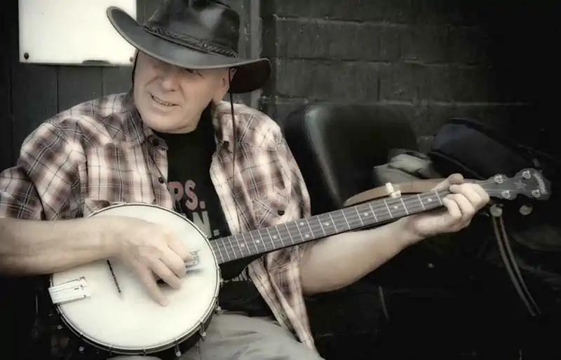 Musician wearing a cowboy hat playing a 5-string banjo outdoors against a dark wall, showing instrument body and neck.
