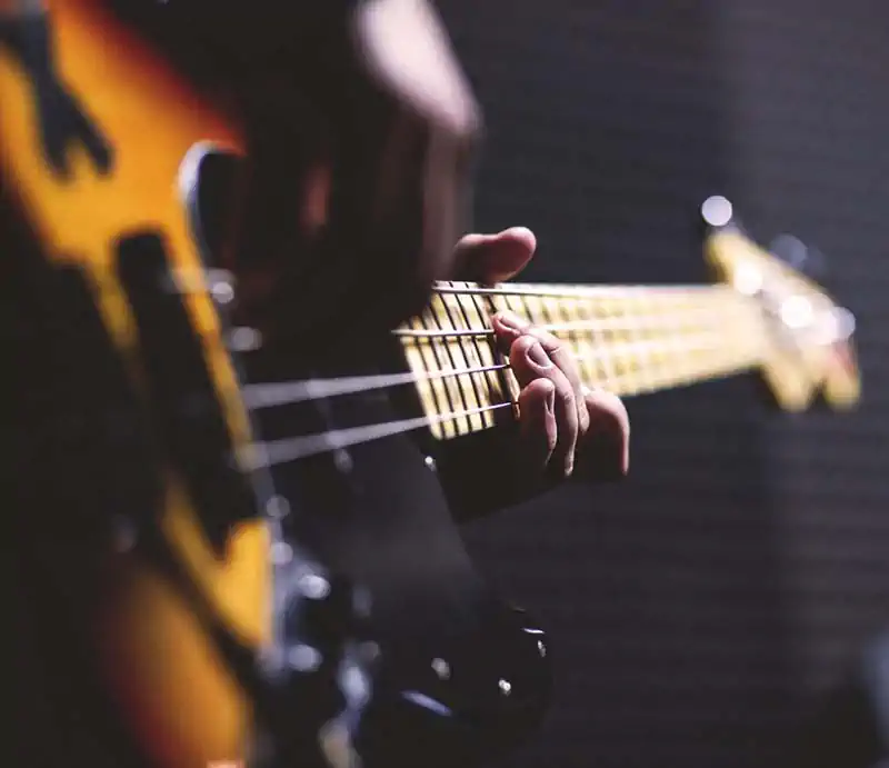 Close-up view of a musician's hand fretting notes on the neck of a sunburst electric bass guitar.