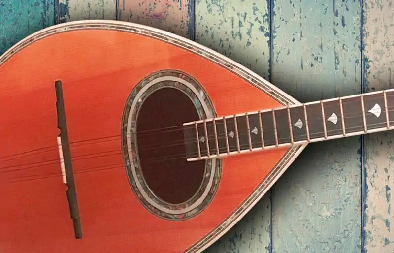 Teardrop-shaped wooden bouzouki body with mother-of-pearl rosette inlay and fretboard resting on rustic painted wood planks.