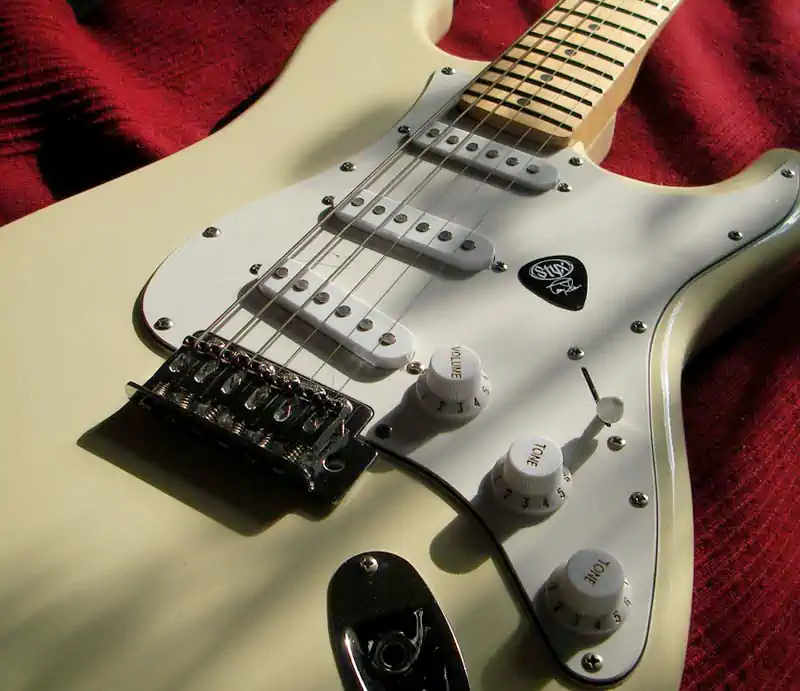 Close-up of a white Fender Stratocaster electric guitar featuring a maple neck, white pickguard, controls, and a black Styx guitar pick.