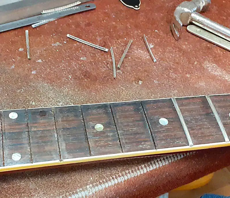 Close-up of a guitar rosewood fretboard undergoing a refret, showing removed fret wire pieces and luthier tools on a workbench.