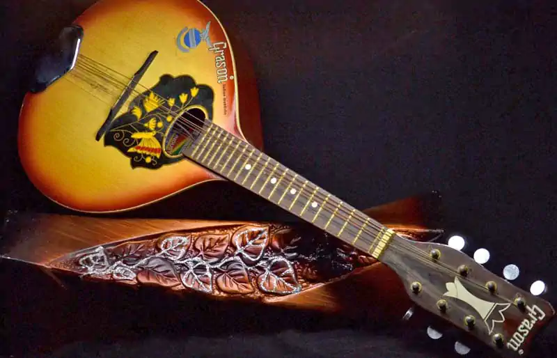 Sunburst Grason acoustic mandolin with decorative floral pickguard resting on a carved wooden base against a dark background.