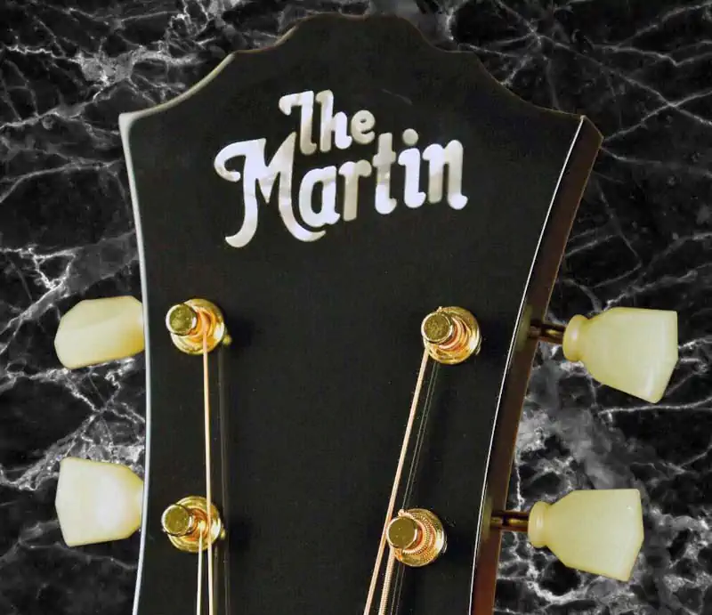 Close-up of a black guitar headstock with mother of pearl The Martin logo inlay, tuning pegs, and gold posts.